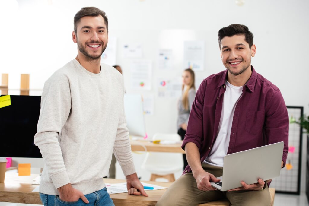 selective focus of smiling marketing managers with laptop leaning on workplace in office